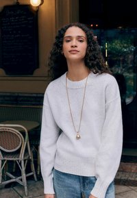 White knit sweater with a round neckline, textured finish, paired with light blue jeans; features a gold necklace with a bell pendant.