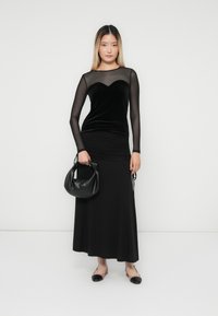 Vrouw staat in een zwart doorzichtig top met lange mouwen en een zwarte maxi rok, terwijl ze een zwarte leren handtas vasthoudt en beige en zwarte platte schoenen draagt.