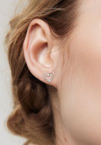 Srebrne kolczyki w kształcie serca, z podwójną linią wzoru, prezentowane na uchu. Tło jest neutralne z widocznymi pasmami włosów.