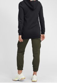 Felpa nera con cappuccio in tessuto morbido, abbinata a pantaloni cargo verde oliva con polsini elasticizzati e tasche laterali. Sneakers bianche con dettagli gialli.