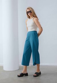 Teal wijdbeenbroek, crèmekleurig aansluitend tanktop en zwarte schuif-sandalen met houten accenten. Glad materiaal, minimalistisch ontwerp en hedendaagse uitstraling.
