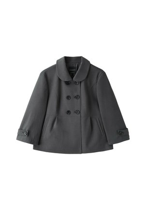 Femme debout, les mains dans les poches, portant une veste grise à double boutonnage, un pantalon large court noir et des ballerines noires sur fond gris.