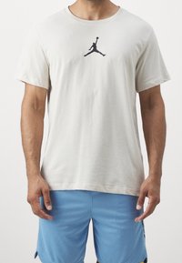 Camiseta de algodón color beige claro con un logo Jumpman negro en el pecho, mangas cortas y un cuello redondo.