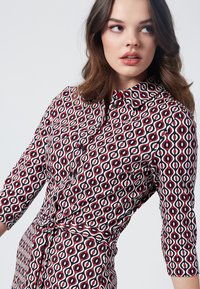 Punainen, musta ja valkoinen kuvioitu mekko, jossa on kaulus, napitus edessä, kolmanneksen mittaiset hihat ja vyötärölle sidottava yksityiskohta. Sujuva kankaan rakenne.