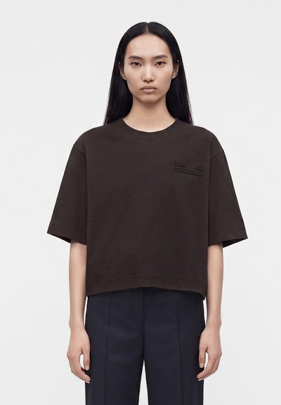 Femme aux longs cheveux noirs et lisses, portant une chemise ample à manches courtes marron foncé et un pantalon foncé, debout devant un fond clair uni.