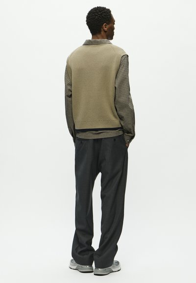 Gilet lavorato a maglia beige sopra una camicia a quadri con bottoni, abbinato a pantaloni grigi larghi e scarpe da ginnastica grigie, visto da dietro.