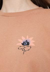 Μπλούζα σε χρώμα σκουριάς με γραφικό λουλούδι που απεικονίζει έναν μαύρο λωτό και σχέδιο από συρματοπλέγματα. Κατασκευασμένη από απαλό βαμβάκι με στρογγυλή λαιμόκοψη.