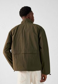 Veste vert olive avec un col haut, coupe ample et tissu texturé. Comprend des manches longues et un ourlet ajustable avec cordon de serrage.