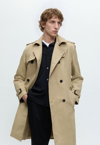 Jeune homme aux cheveux bouclés portant un trench-coat beige sur une chemise noire boutonnée, debout les mains dans les poches devant un fond blanc.