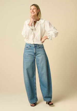 Lachende vrouw staat met hand op kin en heup, draagt een witte blouse met lange mouwen, wijdvallende blauwe jeans, zwarte hakken en een gouden ketting.