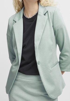 Kvinde iført lysegrøn blazer og nederdel med sort skjorte og sølv snoet kædehalskæde, hænderne bag ryggen.