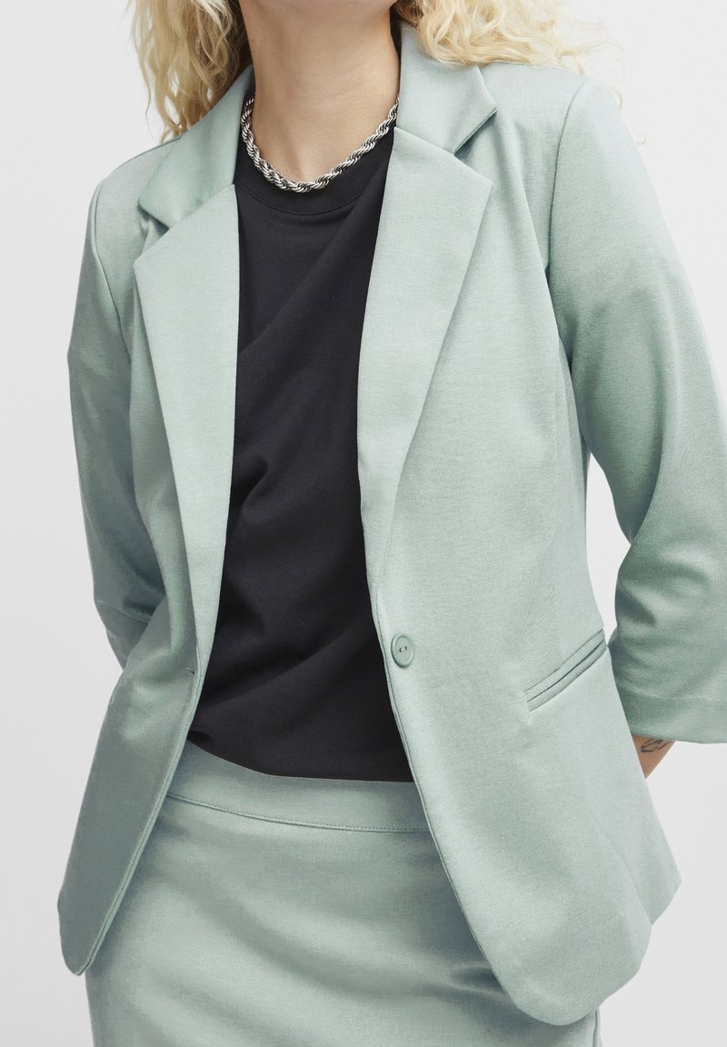 Femme portant un blazer vert clair et une jupe avec une chemise noire et un collier chaîne torsadée argentée, mains derrière le dos.