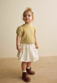 Stickad gul topp med puffärmar som matchas med en vit blommig kjol och bruna ankelstövlar. Har strukturerade mönster och volangdetaljer på strumporna.