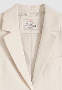 Μπεζ σακάκι Levi Strauss με εγκοπές στο πέτο, ορατή εσωτερική επένδυση και ετικέτα της μάρκας ραμμένη κάτω από το γιακά που υποδεικνύει το μέγεθος μικρό.