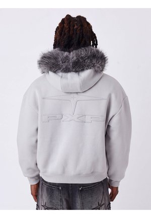 Personne aux cheveux foncés tressés portant un sweat à capuche gris clair avec capuche doublée de fourrure et logo PXP embossé dans le dos, associé à un jean gris foncé.