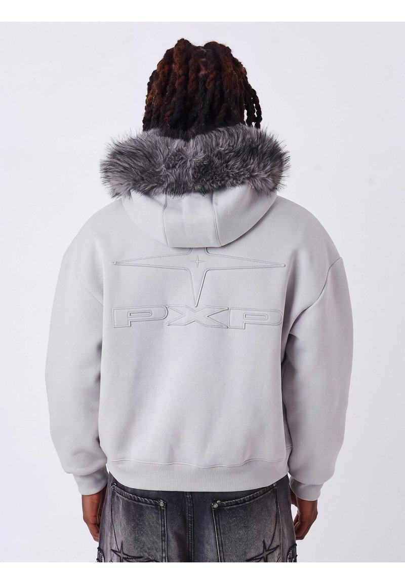 Personne aux cheveux foncés tressés portant un sweat à capuche gris clair avec capuche doublée de fourrure et logo PXP embossé dans le dos, associé à un jean gris foncé.