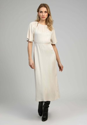 Femme portant une longue robe de couleur crème aux manches courtes volantées et des bottes noires montant jusqu'aux genoux, devant un fond neutre.