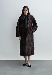 Bordo spalvos dirbtinio kailio paltas su apykakle ir ilgomis rankovėmis, vidutinio ilgio ir lygaus paviršiaus, derinamas su juodais batais.