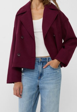Femme portant un manteau court bordeaux à double boutonnage, un débardeur blanc côtelé et un jean bleu clair taille haute, la main dans la poche.