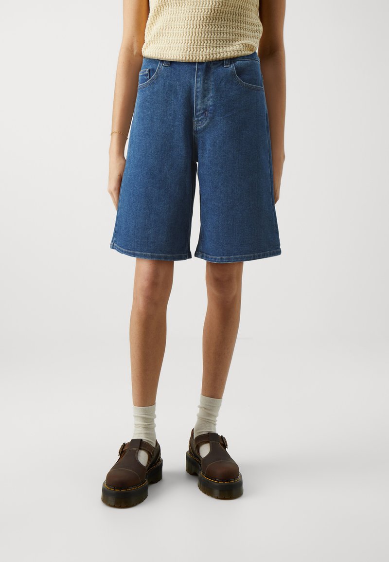 Shorts en denim Bermuda bleu marine, avec cinq poches, une coupe ample et droite. Associés à des chaussures marron et des chaussettes de couleur claire.