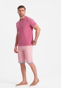 Růžová polo košile s kapsou na hrudi, v kombinaci s vykasanými růžovými šortkami a béžovými slip-on botami. Oblečení má uvolněný střih a jemnou látku.