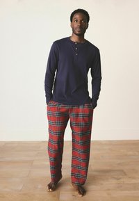 Marineblaues Langarm-Henley-Shirt mit drei Knöpfen, kombiniert mit rot karierten Pyjamahosen mit grünen und schwarzen Linien, vor einem hellen Hintergrund.