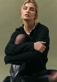 Črn cardigan s gumbom, nosi se preko prosojnih črnih nogavic. Bela perla ogrlica poudarja vratni izrez. Mehka tekstura in sproščen kroj.