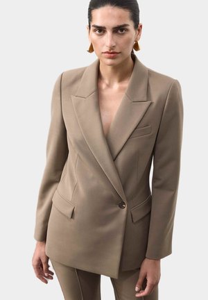 Donna con capelli scuri che indossa orecchini d'oro e un giacca su misura beige abbinata a pantaloni, in piedi davanti a uno sfondo chiaro e semplice.