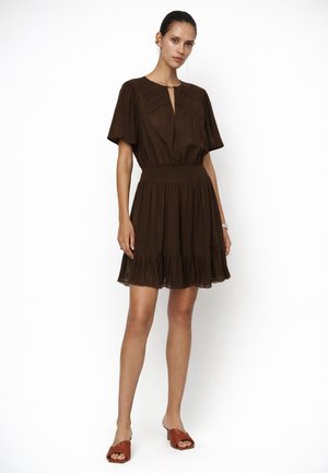 Vrouw staand, gekleed in een korte bruine jurk met een geplooide taille en een gerimpelde zoom, gecombineerd met bruine open teen mule-hakken tegen een witte achtergrond.
