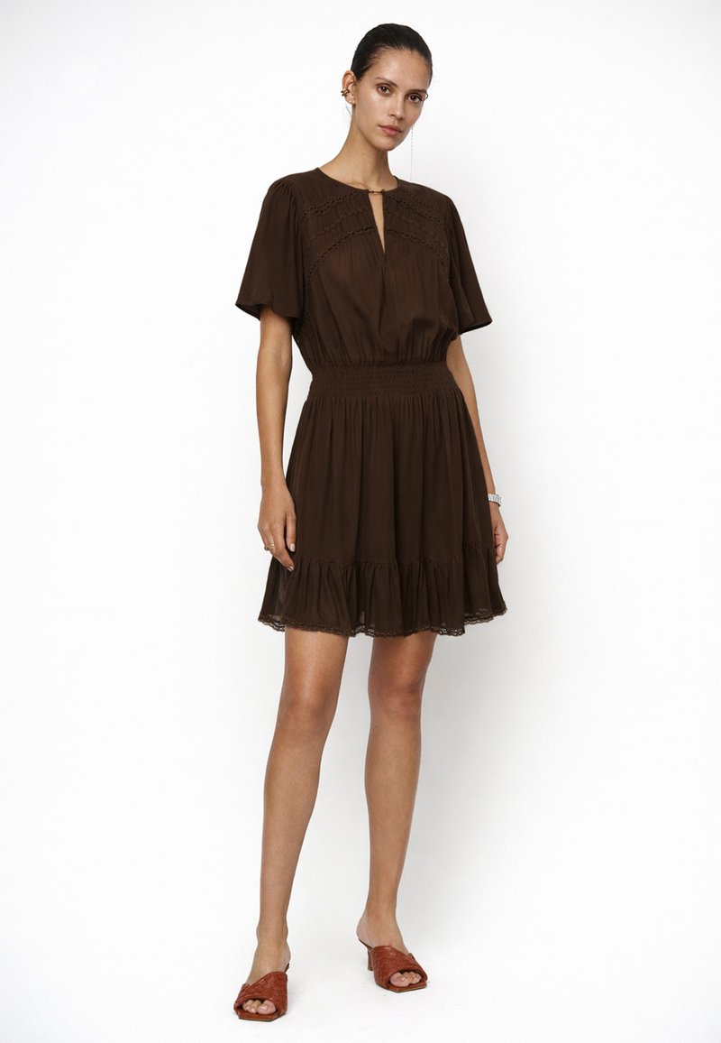 Frau steht und trägt ein kurzes braunes Kleid mit geraffter Taille und Volant-Saum, kombiniert mit braunen offenen Mule-Absätzen vor weißem Hintergrund.