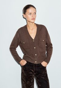 Cardigan lavorato a maglia marrone con scollo a V profondo, due tasche sul petto e bottoni dorati, abbinato a pantaloni di velluto neri. Tessuto liscio e design aderente.