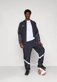 Schwarze Sportjacke und Hose mit weißen Akzenten, vorne mit Reißverschluss und Mesh-Details. Modell steht auf einem Fußball.