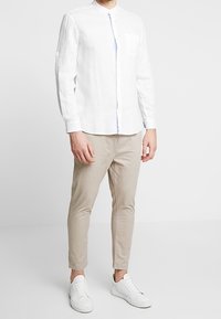 Biała lniana koszula z długim rękawem i kołnierzykiem mandarynka, z jedną kieszenią na piersi. Połączona z beżowymi, dopasowanymi spodniami i białymi trampkami.