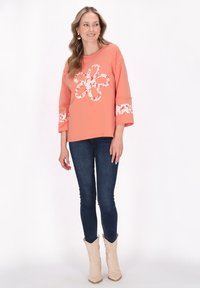 Korallfärgad sweatshirt med blommig applikation, trekvartsärmar med blommigt kantband, parat med mörk denimjeans och ljusa stövlar.