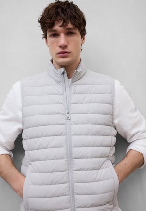 Ung mand står med hænderne i lommerne, iført en lysegrå quiltet vest uden ærmer over en hvid langærmet skjorte mod en ensfarvet baggrund.