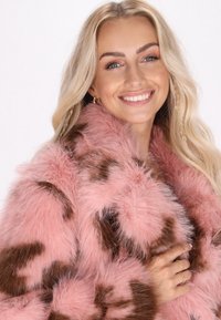Chaqueta de piel sintética en rosa con manchas marrones; textura suave; presenta un cuello alto y solapas anchas; estilizada con anillos en capas en la mano del modelo.