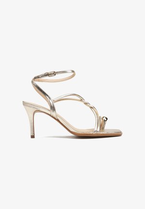 Złote metaliczne sandały na cienkim pasku wokół kostki, z plecionymi paskami z przodu, niskim obcasem i otwartym noskiem zdobionym sercowatą ozdobą.