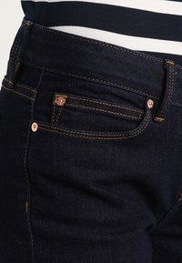 Jeans de mezclilla azul oscuro con remaches de cobre, costuras naranjas y diseño de cinco bolsillos. Textura suave y ajuste entallado en la cintura.
