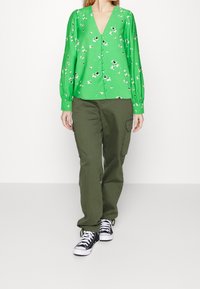 Blusa verde floreale con bottoni, maniche lunghe, scollatura a V e spalle arricciate, abbinata a pantaloni cargo oliva e sneakers nere.