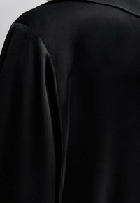 Czarny płaszcz o gładkim, matowym materiale z wyraźnym kołnierzem i wyraźnymi szwami, prezentujący minimalistyczny, dopasowany krój.