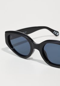 Óculos de sol pretos com um acabamento brilhante, formato de gatinho oversized, lentes azul-escuras e hastes finas com detalhes de marca discretos.