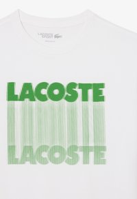 Camiseta deportiva blanca de Lacoste con "LACOSTE" en letras verdes en negrita, distorsionada con líneas verticales verdes debajo del texto.
