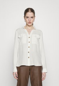 White long-sleeve shirt with a button-up front, two chest pockets, and tortoiseshell buttons. Smooth fabric with a relaxed fit.