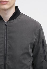 Šedá bomber bunda se zipem, černým žebrovaným límcem a kapsou na zip na rukávu. Hladká textura, minimalistický design.