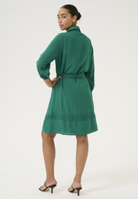 Grön klänning med insnörd midja och krage, med l�ånga ärmar, en texturerad nederkant och slät tyg. Bärs med svarta klackar.
