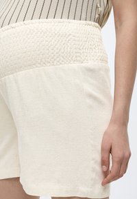 Κρεμ σόρτς με υφή και ψηλή μέση, φορεμένα με αμάνικο μπεζ τοπ με λεπτές μαύρες ρίγες, το χέρι χαλαρά δίπλα στο σώμα, σε λευκό φόντο.