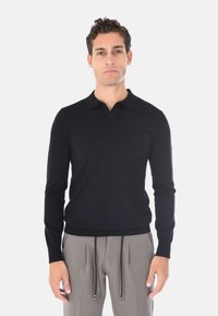 Pullover polo a maniche lunghe di colore nero con scollo a V, polsini e orlo a coste, realizzato in un tessuto liscio e leggero. Abbinato a pantaloni grigi con cordoncino.