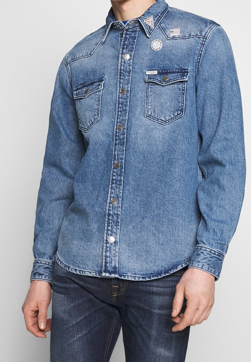 Sininen denim-paita, jossa on kaksi rintataskua, napitus ja hopeiset yksityiskohdat. Sisältää haalistuneen pesun ja erottuvan ommeltavan sauman.