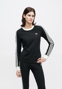 Žena stojí s rukou na boku, oblečená v čiernom teplákovom súprave Adidas s bielymi pruhmi na rukávoch a nohaviciach na jednoduchom bielom pozadí.