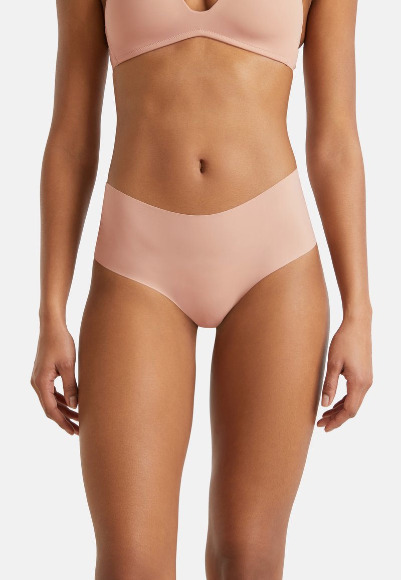Femme portant un ensemble de lingerie nude sans couture, montrant la taille, les hanches et le haut des cuisses sur fond blanc.